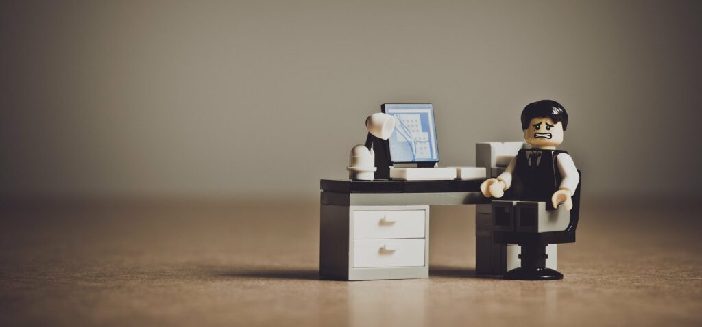 a macro shot of a LEGO figurine sitting at a LEGO office desk and computer. The figurine is facing the camera and wears a worried expression.