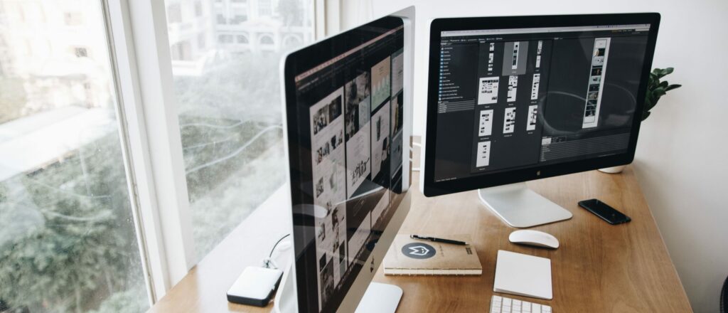 A photograph of two monitors on a wooden desk overlooking a window. A keyboard, mouse, cellphone, and notebook rest in front of the monitors.