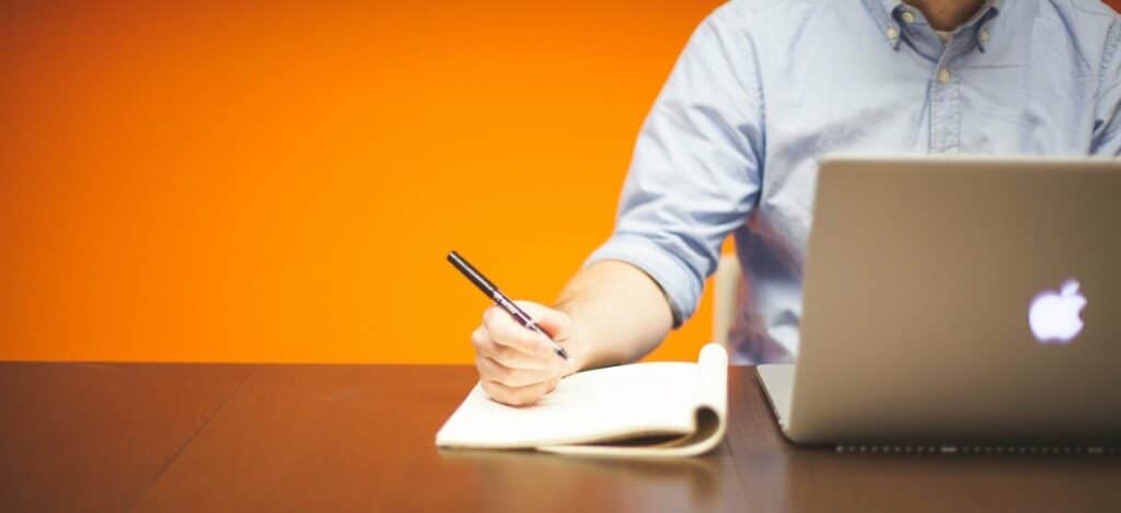 A photograph of a man taking notes in a notebook while working on a laptop.
