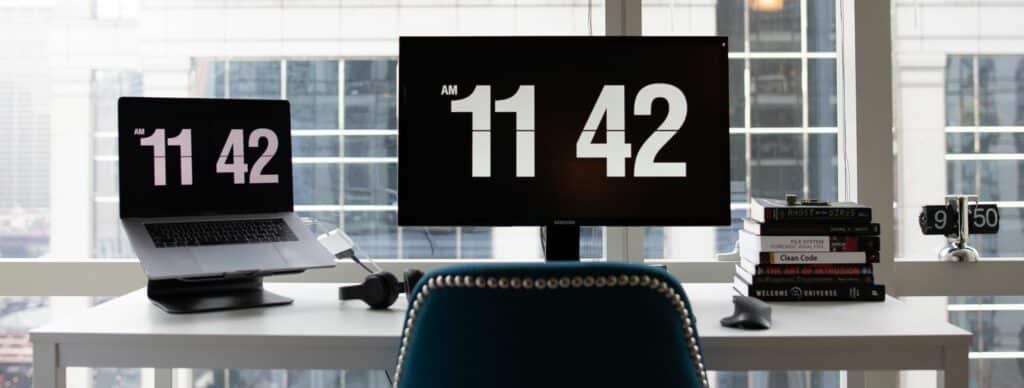 A photograph of an office desk with a laptop and computer monitor. The time is displayed on both screens.