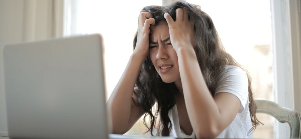 A photograph of a woman holding her head in her hands and frowning at her laptop.
