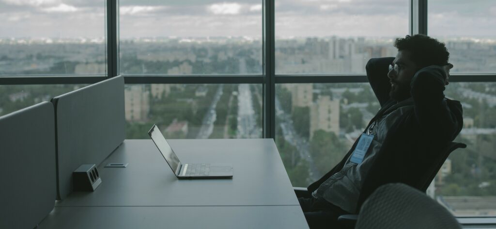 A man in an office overlooking a city skyline works on his laptop.