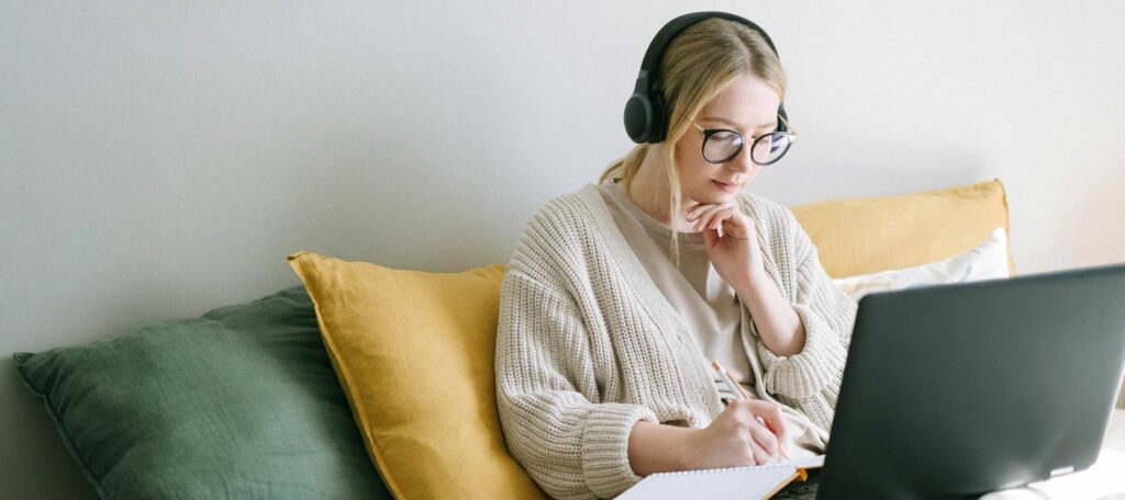 A young woman wear headphones and works on her laptop while sitting on a sofa.