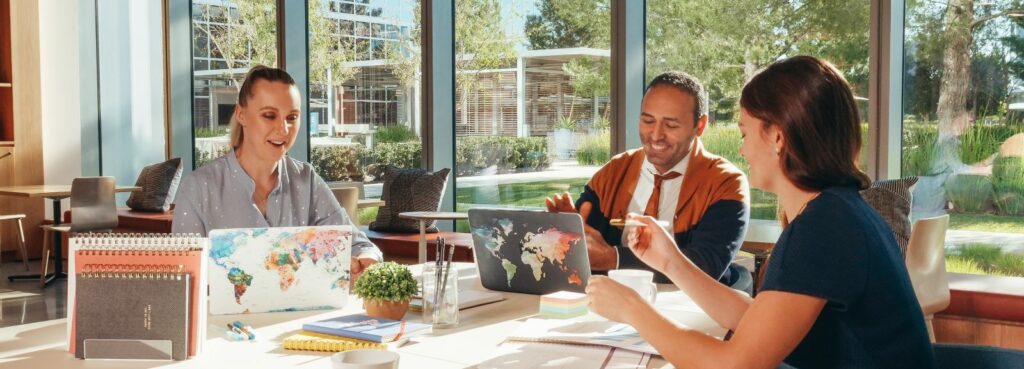 A photograph of a man and two women all working on laptops and smiling as they sit around a table together.