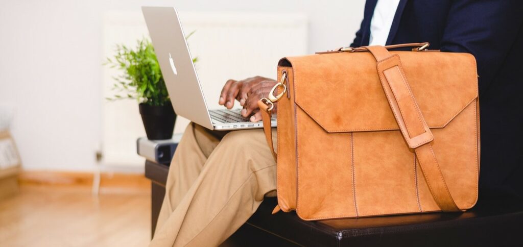 A photograph of a man sitting on a bench and typing on a laptop. A leather briefcase sits beside him.