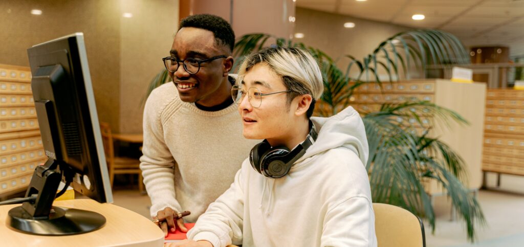 A photograph of two men working on a computer together in a library
