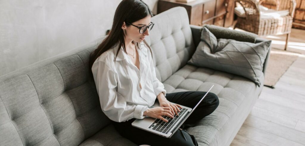 A woman sits cross-legged on a couch while typing on a laptop.
