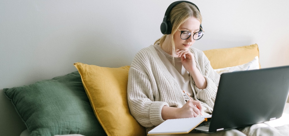 A woman sits on a couch wearing headphones and working on a laptop