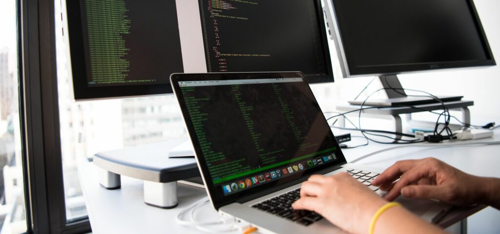 Someone types on a laptop with three additional computer monitors also displayed on their desk. 